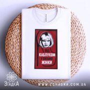 Футболка напис під каблуком, біла, складена на плетеному кошику, фон білий, принт червоний з чорною обводкою, на якому зображена жінка з коротким волоссям та написом 'ПІД КАБЛУКОМ У СИЛЬНОЇ ЖІНКИ'.