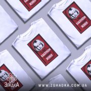 Футболка напис під каблуком, біла, складена на столі, фон сірий, декілька футболок в одному кадрі, принт червоний з чорною обводкою, на якому зображена жінка з коротким волоссям та написом 'ПІД КАБЛУКОМ У СИЛЬНОЇ ЖІНКИ'.