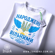 Футболка народжені вільними подарунок. Біла футболка, складена на столі, з принтом у вигляді тризуба та крил, оформленим у блакитних та жовтих кольорах, з етикеткою на зеленій стрічці.