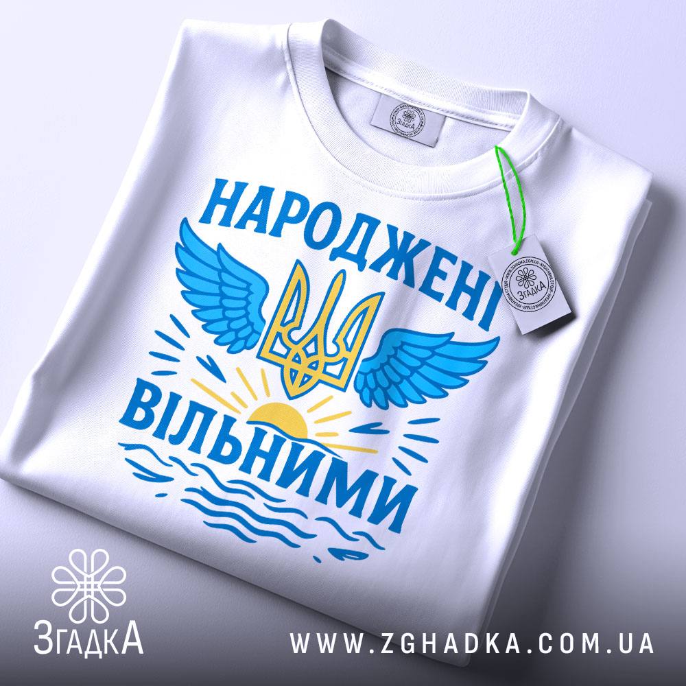 Футболка народжені вільними подарунок. Біла футболка, складена на столі, з принтом у вигляді тризуба та крил, оформленим у блакитних та жовтих кольорах, з етикеткою на зеленій стрічці.
