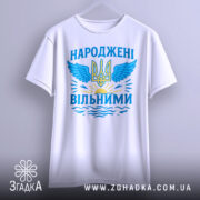 Футболка народжені вільними подарунок. Біла футболка, підвішена на вішалці, з принтом у вигляді тризуба та крил, оформленим у блакитних та жовтих кольорах, на фоні світло-сірого градієнту.