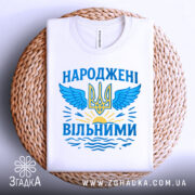 Футболка народжені вільними подарунок. Біла футболка, складена на кошику, з принтом у вигляді тризуба та крил, оформленим у блакитних та жовтих кольорах, на фоні плетеного кошика.