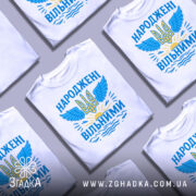 Футболка народжені вільними подарунок. Білі футболки, складені в хаотичному порядку, з принтом у вигляді тризуба та крил, оформленим у блакитних та жовтих кольорах, на фоні світло-сірого градієнту.