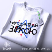 Футболка народжений зіркою подарунок, біла, складена на столі, фон світлий, принт з текстом "НАРОДЖЕНИЙ ЗІРКОЮ" з червоними і синіми зірками, на футболці є етикетка.