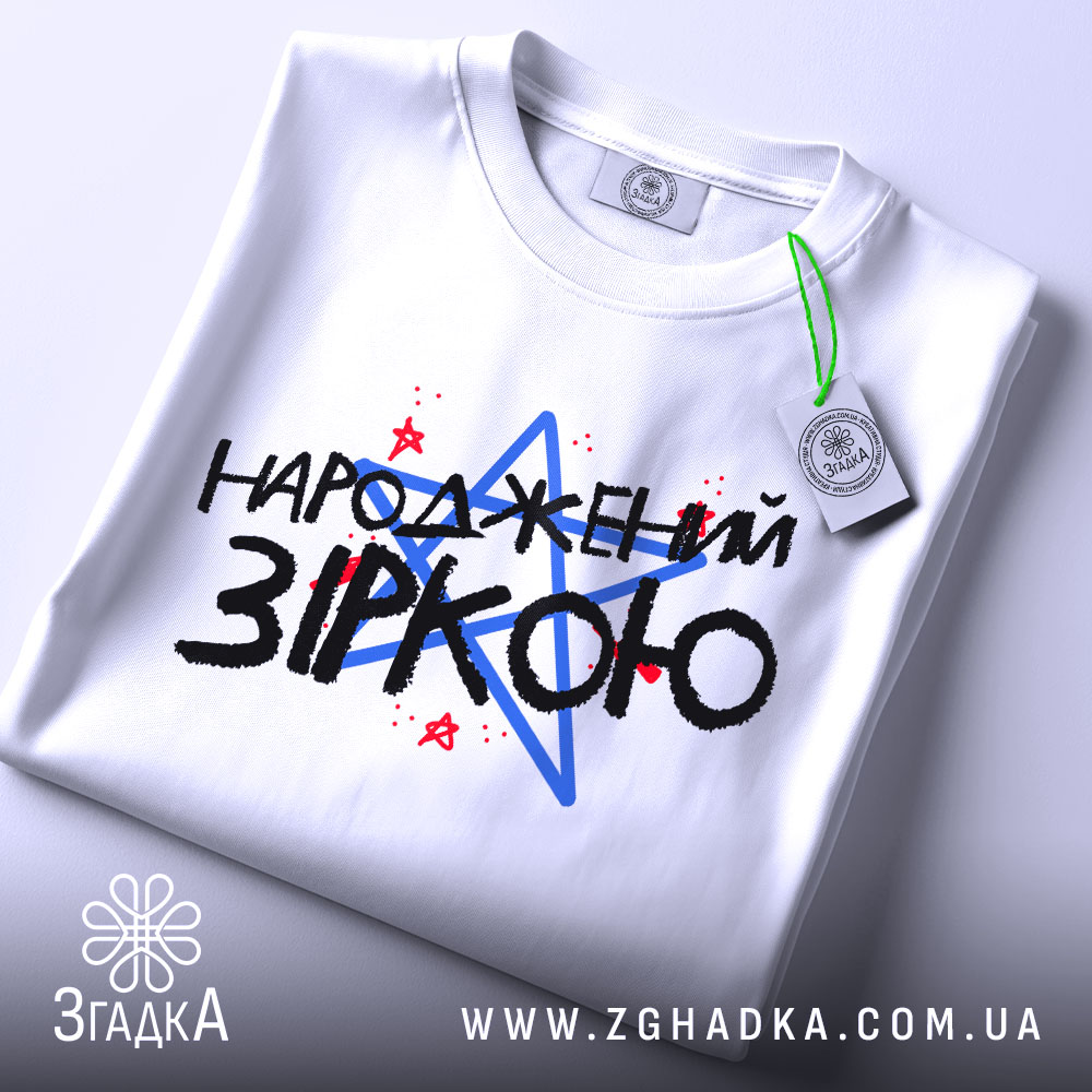 Футболка народжений зіркою подарунок, біла, складена на столі, фон світлий, принт з текстом "НАРОДЖЕНИЙ ЗІРКОЮ" з червоними і синіми зірками, на футболці є етикетка.