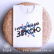 Футболка народжений зіркою подарунок, біла, складена на плетеній кошику, фон світлий, принт з текстом "НАРОДЖЕНИЙ ЗІРКОЮ" з червоними і синіми зірками.