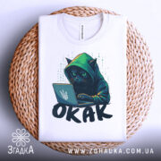 Футболка окак мем подарунок, біла футболка, складена на плетеній кошику, з принтом чорного кота в капюшоні, який працює за ноутбуком, фон білий.