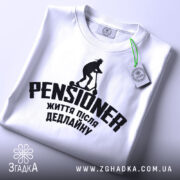 Футболка пенсіонер після дедлайну, біла, розкладена на поверхні, фон світло-сірий, принт чорного кольору з написом 'PENSIONER' та зображенням людини на самокаті, з зеленим ярликом.