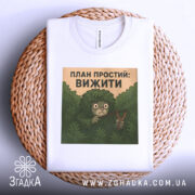 Футболка план простий вижити. Біла футболка, складена на плетеній кошику, з принтом на грудях, фон світло-сірий, принт з персонажем у кущах, що тримає рогатку.