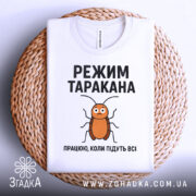 Футболка з принтом таргана, білого кольору, зображена зверху на плетеній кошику, фон світло-сірий, принт з тарганом та текстом "РЕЖИМ ТАРАКАНА ПРАЦЮЮ, КОЛИ ПІДУТЬ ВСІ".