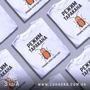 Футболка з принтом таргана, білого кольору, зображена зверху на фоні з кількома футболками, фон світло-сірий, принт з тарганом та текстом "РЕЖИМ ТАРАКАНА ПРАЦЮЮ, КОЛИ ПІДУТЬ ВСІ".