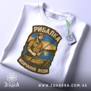 Футболка рибалка на воді, біла, складена на столі, фон білий, принт з рибалкою на човні з вудкою, прикріплений етикет.
