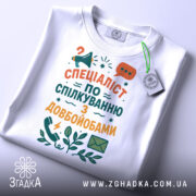Футболка спілкування з довбойобами, білого кольору, складена на поверхні, фон світло-сірий, принт з текстом "СПЕЦІАЛІСТ ПО СПІЛКУВАННЮ З ДОВБОЙОБАМИ" в яскравих кольорах, з елементами мегафона та листа, на футболці є етикетка.