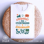 Футболка спілкування з довбойобами, білого кольору, складена на кошику, фон світлий, принт з текстом "СПЕЦІАЛІСТ ПО СПІЛКУВАННЮ З ДОВБОЙОБАМИ" в яскравих кольорах, з елементами мегафона та листа.