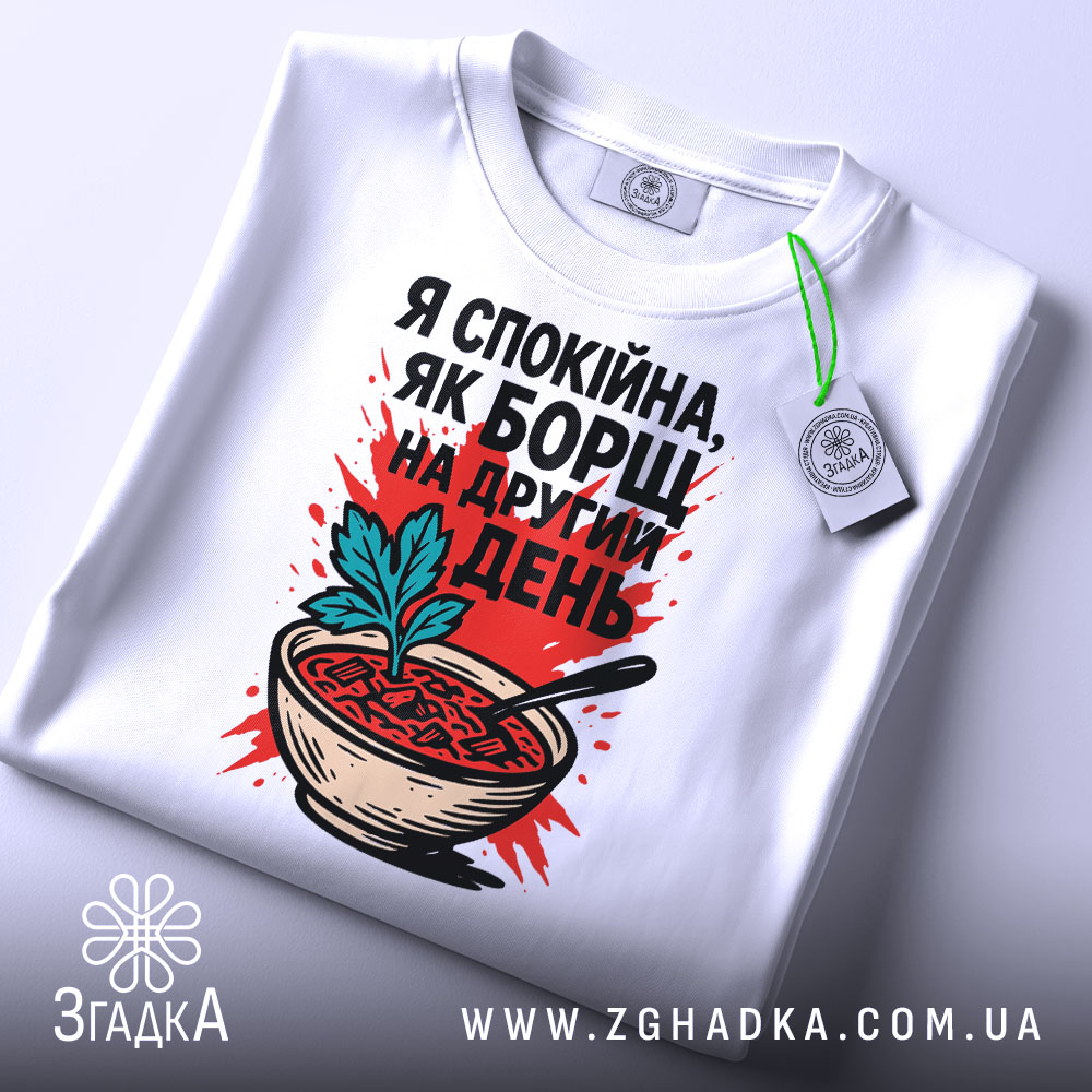 Футболка спокійна як борщ. Біла футболка на столі з видимим етикеткою, принт на грудях, що зображає борщ з зеленню на червоному фоні, злегка згорнута.