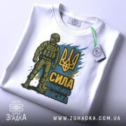 Футболка Сила українського війська, біла, розкладена на поверхні, фон світло-сірий, принт з воїном у формі та тризубом, присутня етикетка з зеленим кантом.