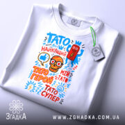 Футболка Тато супер герой, біла, складена на столі, фон світло-сірий, принт з яскравими кольоровими малюнками і текстом, прикріплений етикет.