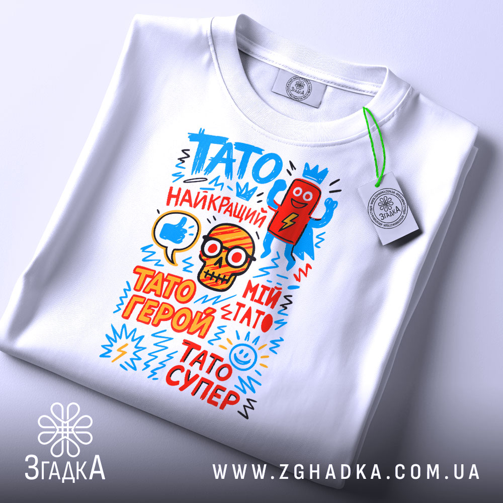 Футболка Тато супер герой, біла, складена на столі, фон світло-сірий, принт з яскравими кольоровими малюнками і текстом, прикріплений етикет.
