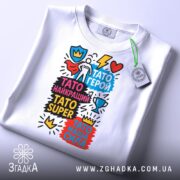 Футболка Тато супер герой, біла, складена на поверхні, фон світлий, принт яскравий з різнокольоровими написами та елементами, на футболці є етикетка.