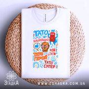 Футболка Тато супер герой, біла, складена на плетеній кошику, фон світлий, принт яскравий з малюнками і текстом.