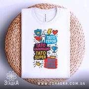 Футболка Тато супер герой, біла, складена на кошику з ротанга, фон світлий, принт яскравий з різнокольоровими написами та графічними елементами.