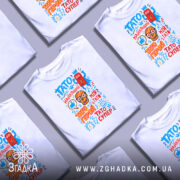 Футболка Тато супер герой, біла, складена на столі, фон світло-сірий, принт з кольоровими елементами, футболки розташовані в хаотичному порядку.