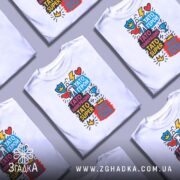 Футболка Тато супер герой, біла, складена на поверхні з декількома іншими футболками, фон сірий, принт яскравий з різнокольоровими написами.