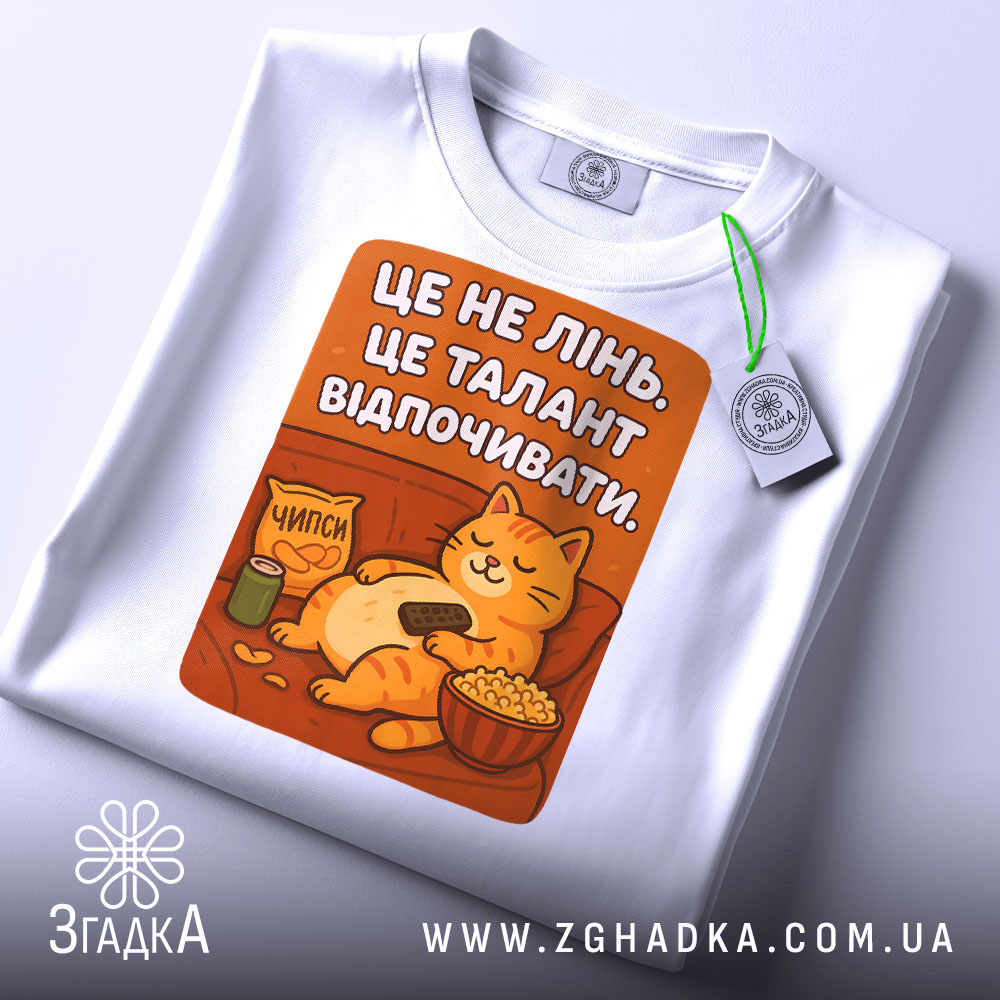 Футболка це не лінь. Біла футболка, складена на поверхні, з принтом кота, що відпочиває. На футболці є етикетка з зеленим шнурком, принт оранжевого кольору.