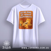 Футболка це не лінь. Біла футболка, підвішена на вішалці, з принтом кота, що відпочиває на дивані. Принт яскравий, з текстом на фоні оранжевого кольору.