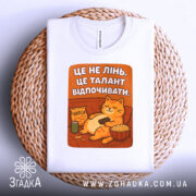 Футболка це не лінь. Біла футболка, складена на кошику, з принтом кота, що відпочиває. Принт оранжевого кольору з текстом, фон світлий.