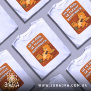 Футболка це не лінь. Біла футболка, розкладена на поверхні, з кількома іншими футболками. Принт кота, що відпочиває, яскравий, з текстом на фоні оранжевого кольору.