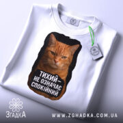 Футболка тихий не спокійний, біла, з верхнього ракурсу, на столі, з принтом оранжевого кота, текстом чорного кольору та етикеткою.