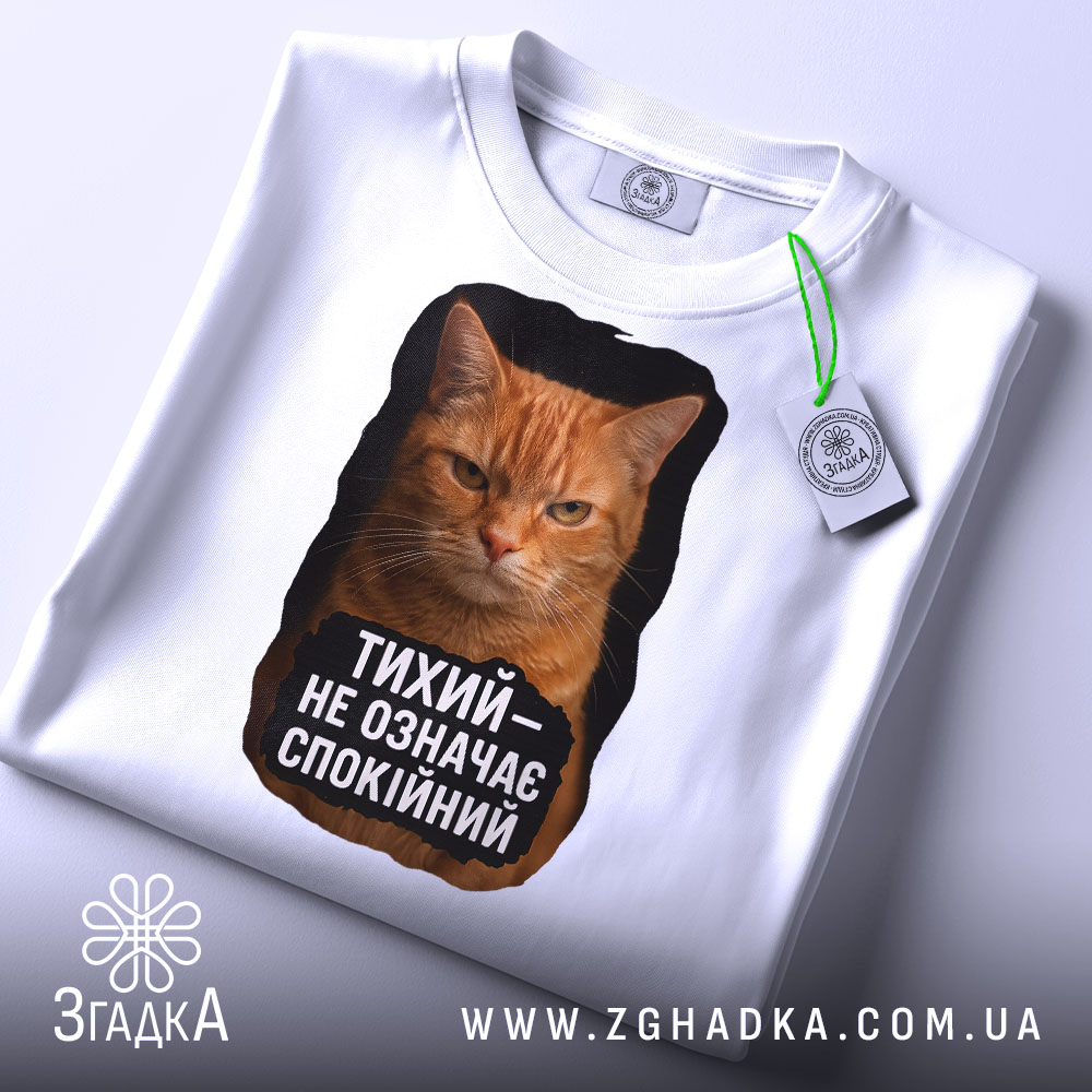 Футболка тихий не спокійний, біла, з верхнього ракурсу, на столі, з принтом оранжевого кота, текстом чорного кольору та етикеткою.