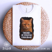 Футболка тихий не спокійний, біла, з верхнього ракурсу, на плетеній кошику, з принтом оранжевого кота та текстом чорного кольору.