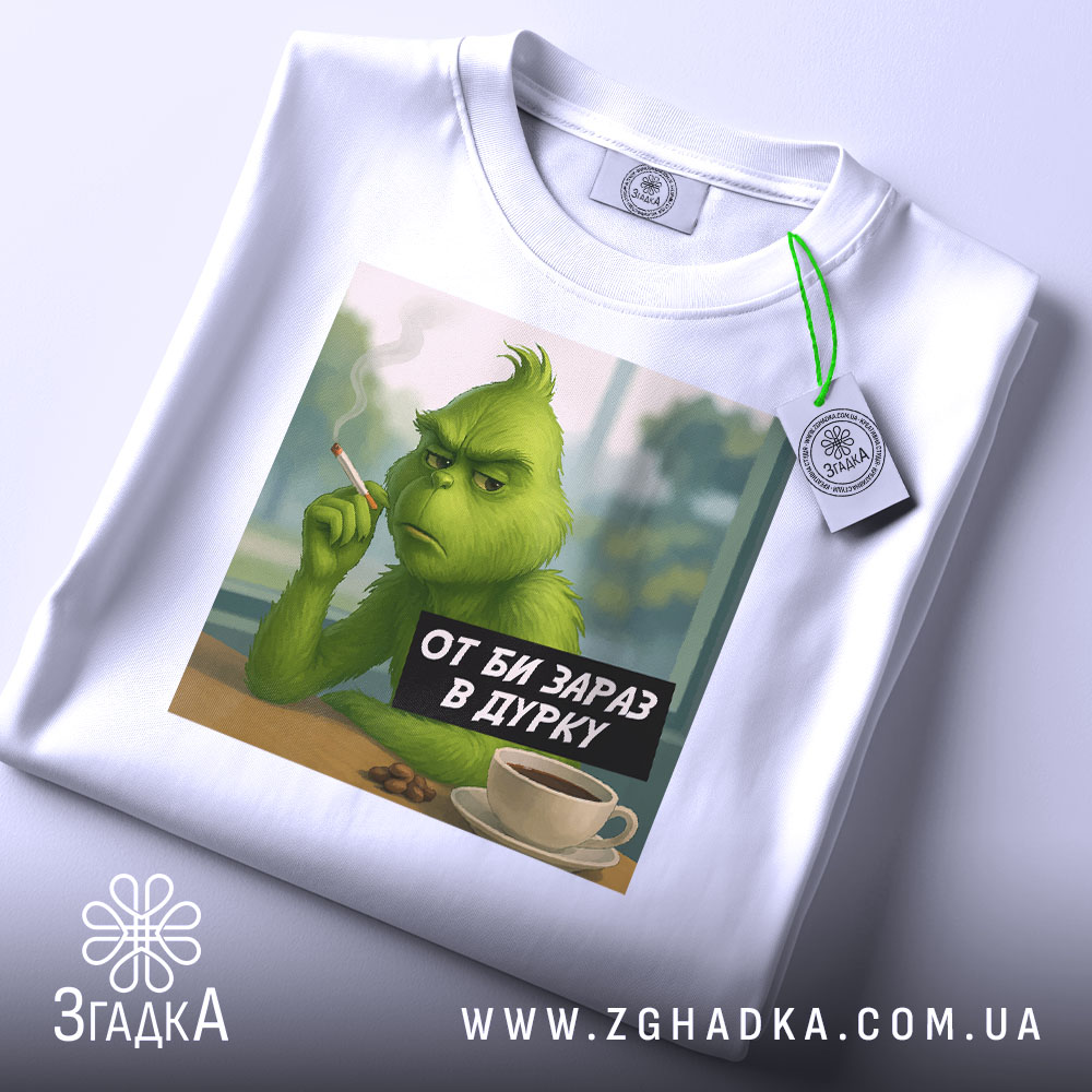 Футболка в дурку подарунок, біла футболка складена, принт з зеленим персонажем, що курить, на фоні видно вікно, деталі принта чіткі, на футболці прикріплений етикет.