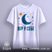 Футболка Вір у себе, біла, на вішалці, принт з місяцем, зірками та кометою, фон сірий.