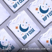 Футболка Вір у себе, біла, на столі, кілька футболок з принтом з місяцем, зірками та кометою, фон сірий.