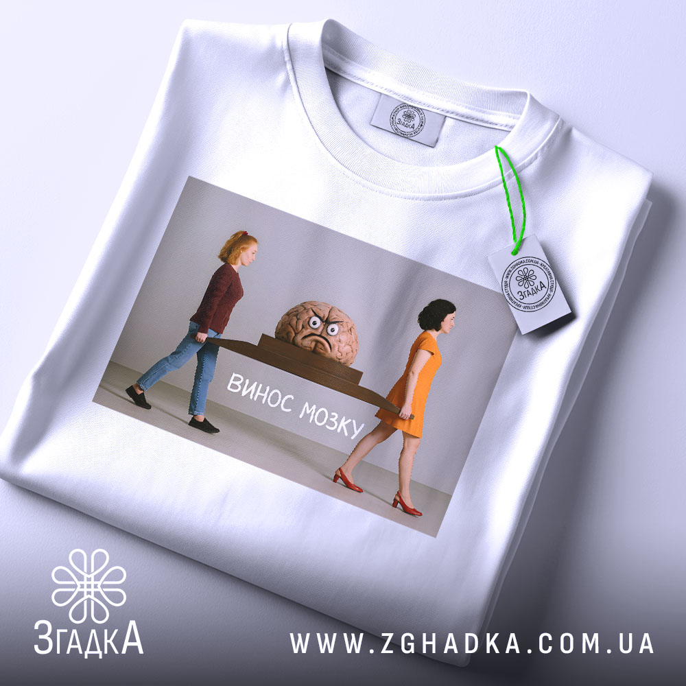 Футболка винос мозку подарунок, біла футболка складена на поверхні, ракурс зверху, фон світло-сірий, принт з двома жінками, які несуть мозок на столі, текст 'ВИНОС МОЗКУ' під малюнком, на футболці є етикетка.