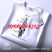 Футболка з автомобільним дизайном, біла, складена на столі, фон світлий, принт з малюнком свічки запалювання та текстом "Гаражний культ" червоного кольору, з етикеткою.