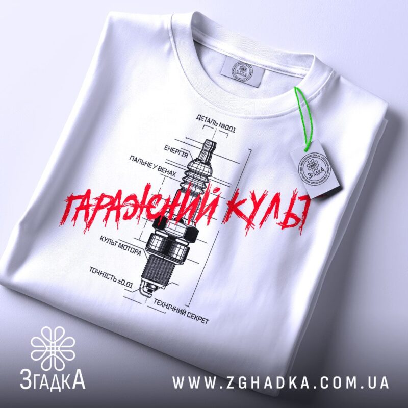 Футболка з автомобільним дизайном, біла, складена на столі, фон світлий, принт з малюнком свічки запалювання та текстом "Гаражний культ" червоного кольору, з етикеткою.