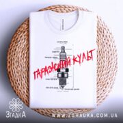 Футболка з автомобільним дизайном, біла, складена на кошику, фон світлий, принт з малюнком свічки запалювання та текстом "Гаражний культ" червоного кольору.