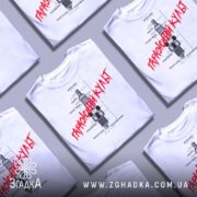 Футболка з автомобільним дизайном, біла, розкладена на столі, фон світло-сірий, кілька футболок з принтом "Гаражний культ" червоного кольору.