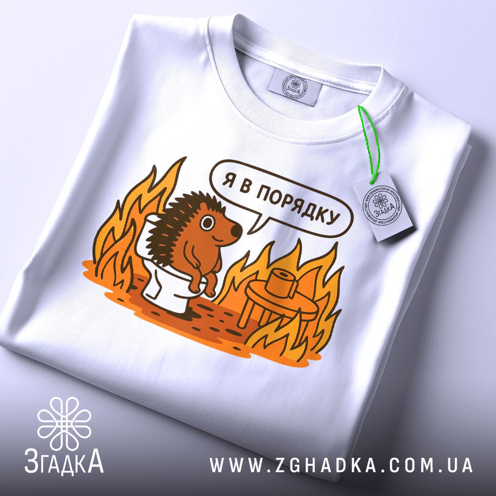 Футболка з їжаком в порядку. Біла футболка, складена на поверхні, з принтом їжака на унітазі. Принт яскравий, з оранжевими вогнями навколо. На футболці є етикетка з зеленим шнурком. Фон світлий, з легким градієнтом.