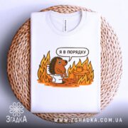 Футболка з їжаком в порядку. Біла футболка, складена на плетеній основі, з принтом їжака на унітазі. Принт яскравий, з оранжевими вогнями. Фон світлий, з плетеним текстурованим елементом.