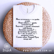 Футболка з кумедними написами, білого кольору, складена на плетеній тарілці, фон білий, принт чорного кольору з текстом на українській мові.