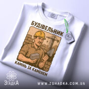 Футболка з малюнком будівельника, білого кольору, зображена з верхнього ракурсу, фон світлий, принт з будівельником у жовтому касці, що тримає камінь і лопату, текст під малюнком: 'Камінь за каменем', на футболці є етикетка.