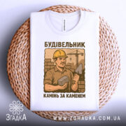 Футболка з малюнком будівельника, білого кольору, зображена на плетеній кошику, фон натуральний, принт з будівельником у жовтому касці, що тримає камінь і лопату, текст під малюнком: 'Камінь за каменем'.