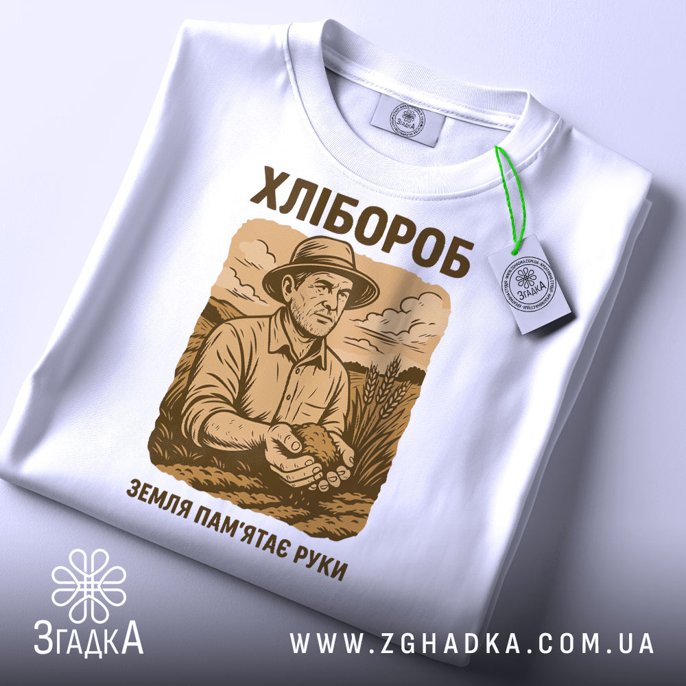 Футболка з малюнком хлібороба, білого кольору, зображена в горизонтальному положенні на білому фоні, принт з малюнком хлібороба в капелюсі, що тримає в руках землю, текст "ХЛІБОРОб" та "ЗЕМЛЯ ПАМ'ЯТАЄ РУКИ" в коричневих тонах, на комірці є бірка.