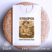 Футболка з малюнком хлібороба, білого кольору, зображена в горизонтальному положенні на плетеному кошику, фон білий, принт з малюнком хлібороба в капелюсі, що тримає в руках землю, текст "ХЛІБОРОб" та "ЗЕМЛЯ ПАМ'ЯТАЄ РУКИ" в коричневих тонах.