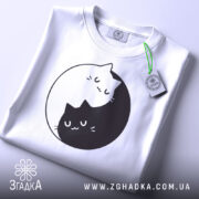 Футболка з малюнком котів білого кольору, складена на поверхні, з видимим принтом двох котів, один з яких чорний, а інший білий, що обіймаються в кругу. На футболці є етикетка зеленого кольору.